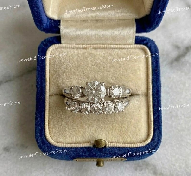 May include: A set of two diamond rings in a blue velvet ring box. The top ring has a large, round center stone flanked by two smaller stones. The bottom ring has a row of smaller diamonds. The box is open.