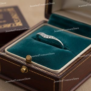 K&ouml;nnte beinhalten: Ein silberfarbener Ring mit geschwungenem Design, besetzt mit kleinen, funkelnden Steinen. Der Ring liegt in einer offenen, dunkelbraunen Schmuckschatulle mit teal-farbenem Samt. Die Schatulle ist aufwendig verziert.