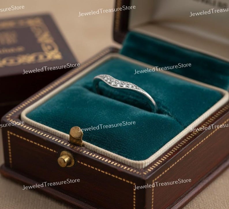 K&ouml;nnte beinhalten: Ein silberfarbener Ring mit geschwungenem Design, besetzt mit kleinen, funkelnden Steinen. Der Ring liegt in einer offenen, dunkelbraunen Schmuckschatulle mit teal-farbenem Samt. Die Schatulle ist aufwendig verziert.