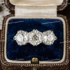 Op de afbeelding: Een zilveren ring met drie grote, rond geslepen diamanten. De ring wordt gepresenteerd in een vintage sieradendoosje met blauwe fluwelen voering en een goudkleurige sluiting en afwerking.