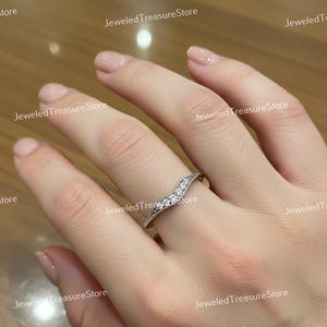 K&ouml;nnte beinhalten: Ein silberfarbener Ring mit geschwungenem Design, besetzt mit kleinen, klaren Edelsteinen. Der Ring befindet sich an einem Finger und zeigt seine filigranen Details und funkelnden Steine. Das Band des Rings hat einen strukturierten Rand.