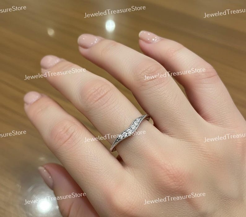 K&ouml;nnte beinhalten: Ein silberfarbener Ring mit geschwungenem Design, besetzt mit kleinen, klaren Edelsteinen. Der Ring befindet sich an einem Finger und zeigt seine filigranen Details und funkelnden Steine. Das Band des Rings hat einen strukturierten Rand.