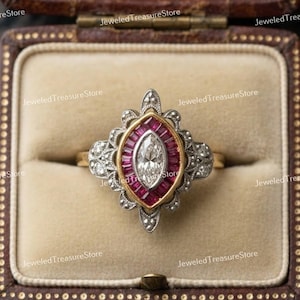 May include: A vintage-style ring with a central oval-shaped diamond, surrounded by a halo of small, rectangular rubies. The ring has a gold band and ornate silver detailing, set in a brown jewelry box.