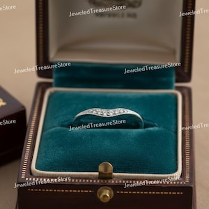 K&ouml;nnte beinhalten: Ein silberner Ring mit geschwungenem Design, besetzt mit kleinen, klaren Edelsteinen. Der Ring wird in einer dunkelbraunen, samtgef&uuml;tterten Schmuckschatulle mit teal-farbenem Innenraum pr&auml;sentiert. Die Schatulle ist geschlossen.