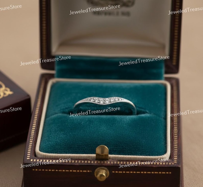 K&ouml;nnte beinhalten: Ein silberner Ring mit geschwungenem Design, besetzt mit kleinen, klaren Edelsteinen. Der Ring wird in einer dunkelbraunen, samtgef&uuml;tterten Schmuckschatulle mit teal-farbenem Innenraum pr&auml;sentiert. Die Schatulle ist geschlossen.