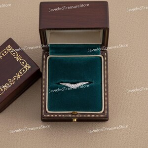 K&ouml;nnte beinhalten: Ein silberner Ring mit geschwungenem Design, besetzt mit kleinen, funkelnden Steinen, liegt in einer dunkelbraunen Schmuckschatulle mit gr&uuml;nem Samtinterieur. Die Schachtel ist ge&ouml;ffnet und zeigt den Ring. Eine weitere Schachtel ist teilweise sichtbar.
