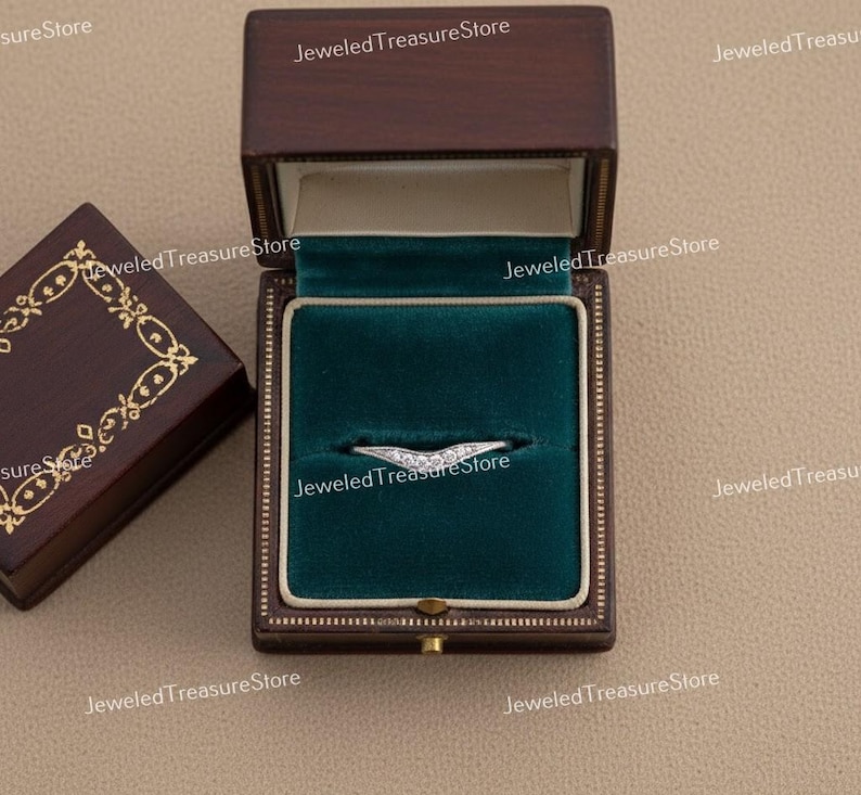 K&ouml;nnte beinhalten: Ein silberner Ring mit geschwungenem Design, besetzt mit kleinen, funkelnden Steinen, liegt in einer dunkelbraunen Schmuckschatulle mit gr&uuml;nem Samtinterieur. Die Schachtel ist ge&ouml;ffnet und zeigt den Ring. Eine weitere Schachtel ist teilweise sichtbar.