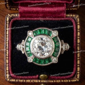 May include: An antique-style ring with a large, clear, round-cut central stone. The ring features a halo of emerald-cut green stones and smaller accent diamonds. The ring is set in a silver-coloured metal and is displayed in a burgundy velvet ring box.