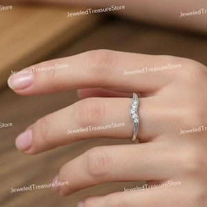K&ouml;nnte beinhalten: Ein silberfarbener Ring mit geschwungenem Design, besetzt mit kleinen, klaren Edelsteinen. Der Ring hat eine Reihe von Steinen und zarte Details entlang des Bandes. Der Ring befindet sich am Finger einer Person.