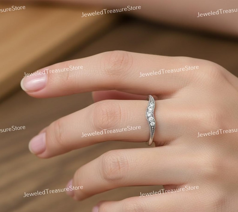 K&ouml;nnte beinhalten: Ein silberfarbener Ring mit geschwungenem Design, besetzt mit kleinen, klaren Edelsteinen. Der Ring hat eine Reihe von Steinen und zarte Details entlang des Bandes. Der Ring befindet sich am Finger einer Person.