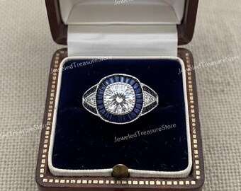 May include: A silver ring featuring a large, round, brilliant-cut clear center stone surrounded by a halo of deep blue baguette-cut stones. The band is accented with small, clear, round stones set in a triangular pattern on each side.