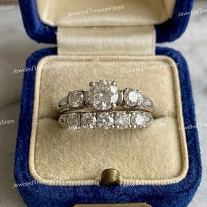 May include: A two-piece diamond ring set in a blue velvet ring box. The engagement ring features a large, round center stone flanked by smaller diamonds. The wedding band has a row of smaller diamonds. The rings are set in a silver metal.