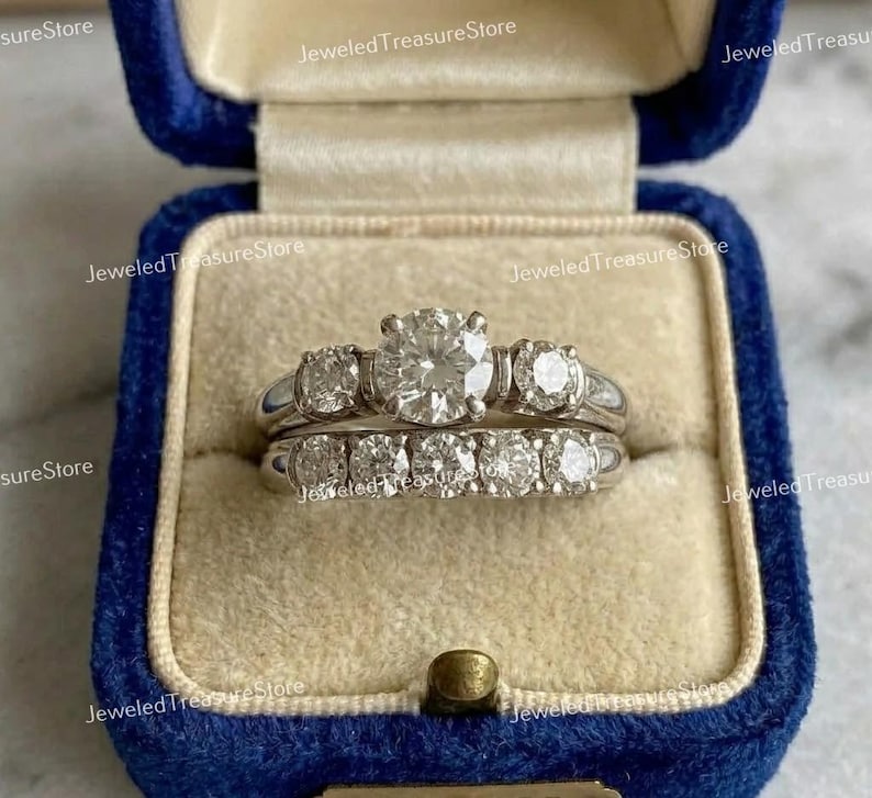 May include: A two-piece diamond ring set in a blue velvet ring box. The engagement ring features a large, round center stone flanked by smaller diamonds. The wedding band has a row of smaller diamonds. The rings are set in a silver metal.