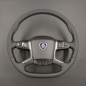 May include: A black steering wheel with a perforated grip and red stitching. The centre features a logo with a blue, red, and white emblem. Control buttons are visible on the wheel's spokes.