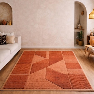 May include: A geometric rug in shades of orange and rust. The rug features a series of rectangular and triangular shapes, creating a modern design. The rug is placed on a wooden floor, adding warmth to the room.