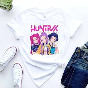 Peut inclure: T-shirt blanc avec un graphisme de trois filles de style anime aux cheveux roses, violets et bleus. Le mot "HUNTRAX" est imprimé au-dessus de l'image en rose. Le t-shirt est associé à un jean bleu et des baskets blanches.