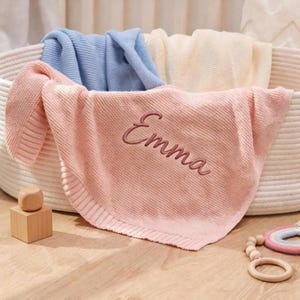 May include: A pink knitted baby blanket embroidered with the name "Emma". The blanket is folded in a white woven basket, with blue and cream blankets visible. A wooden block and a toy are in the foreground. The blanket is suitable for a baby.