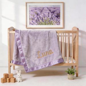 May include: A lavender baby blanket with a satin border and the name "Luna" embroidered in gold. The blanket is draped over a wooden crib. A framed picture of lavender fields hangs above the crib.