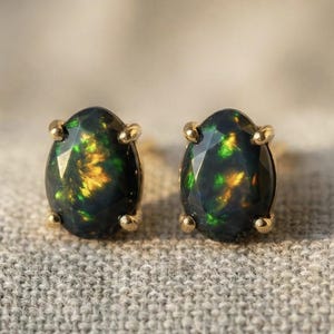 May include: A pair of stud earrings featuring oval-shaped black opals with flashes of green and gold. The opals are set in gold prongs, creating a striking contrast. The earrings are displayed on a textured, neutral-toned surface.