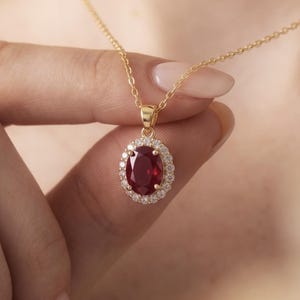 May include: A gold necklace with a pendant. The pendant has a large, oval, deep red gemstone, encircled by smaller, clear gemstones. The chain and pendant setting are gold, creating a classic and elegant design. The necklace is a piece of jewellery.