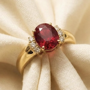 Puede incluir: Un anillo de oro con un gran rubí de corte ovalado. El rubí es de color rojo intenso y está flanqueado por piedras preciosas más pequeñas y transparentes. El anillo está engastado en una banda de oro, un diseño clásico y elegante.