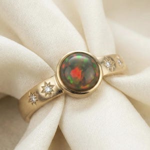 May include: A gold ring featuring a large, round, black opal with red and green flashes. The band is adorned with small starburst designs and inset diamonds, creating a celestial theme. The ring is set against a white fabric.
