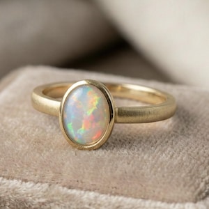 May include: A gold ring with an oval opal gemstone. The opal displays a play of color with flashes of red, green, and yellow. The ring has a simple, elegant design, perfect for everyday wear.