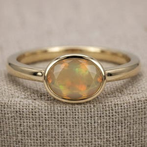 Puede incluir: Anillo de oro con una piedra preciosa de ópalo ovalada. El ópalo muestra tonos de naranja, amarillo y verde. El anillo tiene una banda pulida y un engaste biselado. La piedra preciosa está asegurada en el engaste.