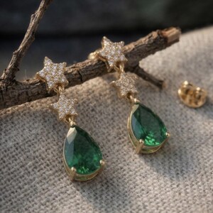 May include: Gold-coloured earrings with a star and teardrop design. The earrings have a star-shaped top encrusted with small, clear stones, and a large, emerald-green teardrop-shaped stone. A gold earring back is also visible.