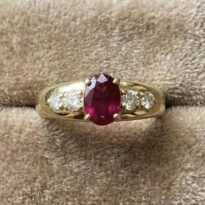 Peut inclure: Une bague en or ornée d'un rubis rouge foncé ovale en son centre, flanqué de trois petites pierres claires de chaque côté. La bague est présentée sur un fond beige doux, mettant en valeur le design et la couleur du bijou.