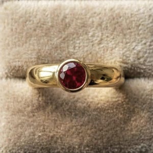 Puede incluir: Un anillo de oro con una piedra preciosa redonda de color rojo intenso engastada en un bisel. El anillo se muestra en un joyero de terciopelo beige, mostrando la banda de oro pulido y el color vibrante de la piedra preciosa.