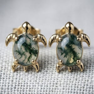 May include: A pair of gold-coloured turtle stud earrings. Each earring features a clear, oval-shaped stone with green and white inclusions, set within a gold-toned turtle frame. The earrings are displayed on a textured, off-white surface.