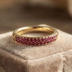 Puede incluir: Un anillo de oro con dos filas de pequeñas piedras preciosas rojas y redondas. El anillo está sobre una superficie beige suave. La banda de oro tiene un acabado pulido. Las piedras preciosas están muy juntas, creando una banda continua.