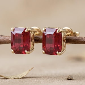 May include: A pair of stud earrings featuring rectangular, emerald-cut red gemstones set in yellow gold. The metal settings display intricate, engraved scrollwork patterns along the sides, and each earring is secured with a gold post backing.