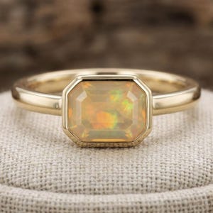 May include: A gold ring featuring a rectangular opal gemstone. The opal displays a warm, iridescent colour palette with flashes of orange, yellow, and green. The ring's band is smooth and polished, and the gemstone is set in a bezel setting.