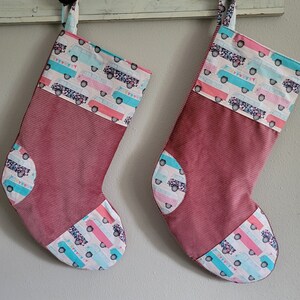 May include: Two Christmas stockings with a burgundy corduroy body and a patterned top and toe. The pattern features colorful vintage vans with floral and banner accents. The stockings are hanging on a white wooden surface.