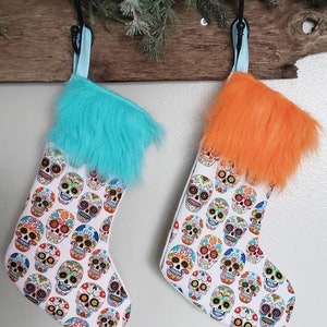 May include: Two Christmas stockings with a white background and a pattern of colorful sugar skulls. One stocking has a turquoise fur cuff and the other has an orange fur cuff.