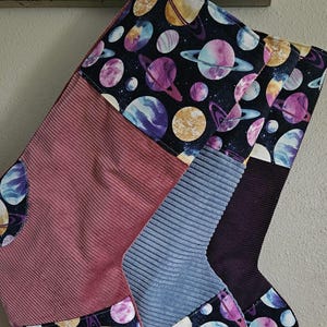 May include: Three Christmas stockings made of corduroy fabric. The stockings are decorated with a patchwork design of different colors and patterns. The top of the stockings features a black fabric with a watercolor print of planets and stars. The bottom of the stockings features a pink, blue, and purple corduroy fabric.