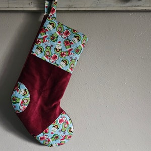 May include: A burgundy Christmas stocking with a blue patterned top and accents. The pattern features avocado characters in Santa hats and festive attire. The stocking has a loop for hanging.