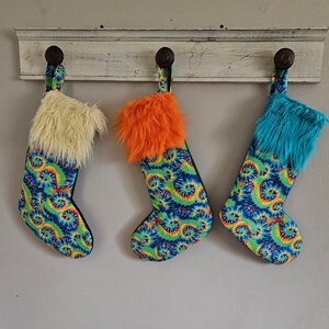 May include: Three Christmas stockings with tie-dye patterns in green, blue, and orange. Each stocking has a fluffy white, orange, or blue trim.