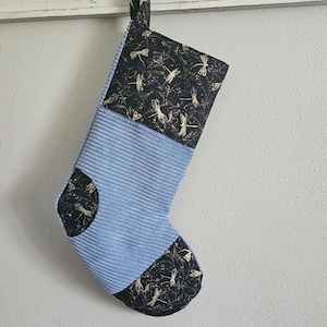 May include: A blue and black Christmas stocking with a dragonfly pattern. The stocking is made of corduroy and has a loop at the top for hanging.