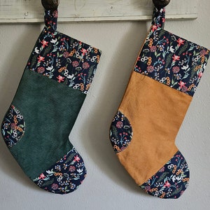 May include: Two Christmas stockings, one green and one tan, hang from a white wooden bar. The stockings have a corduroy texture and are decorated with a floral pattern in shades of blue, red, and white.