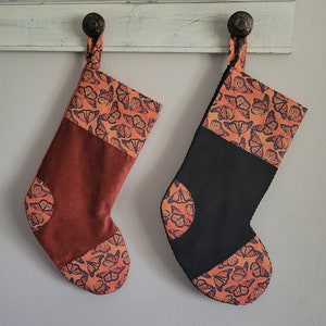 May include: Two Christmas stockings, one rust-colored and one black, hang from a white wooden rack. Both stockings feature a butterfly pattern in orange and black on the cuff and toe. The rust-colored stocking has a corduroy texture.