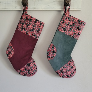 May include: Two Christmas stockings. One is burgundy with a poinsettia pattern on the cuff and toe. The other is teal with the same poinsettia pattern. Both have a loop for hanging.