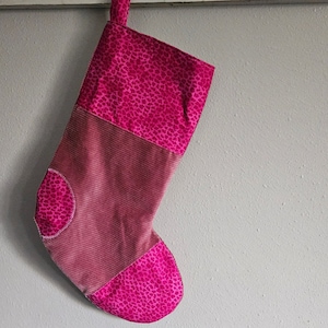 May include: A pink and red Christmas stocking with a leopard print design. The stocking is made of two different fabrics, one with a corduroy texture and the other with a smooth, shiny finish.