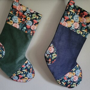 May include: Two Christmas stockings. One is dark green corduroy, the other is navy blue corduroy. Both have a cuff and toe patch with a colorful mushroom and floral pattern. The stockings are hanging against a white wall.