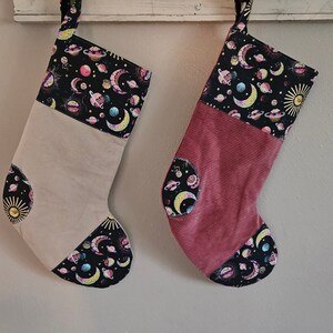 May include: Two Christmas stockings with a celestial theme. One is beige, the other is red. Both have a black top and toe with a colorful space pattern of planets, moons, and stars. The stockings are hanging against a white background.