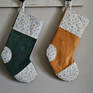 May include: Two handmade Christmas stockings. One is dark green corduroy, the other is golden yellow corduroy. Both have a white floral pattern at the top and on the toe. Each stocking has a fabric loop for hanging.