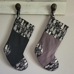 Peut inclure: Deux chaussettes de Noël avec un motif de chat noir et blanc. Une chaussette est noire avec un revers en velours côtelé gris, et l'autre est grise avec un revers en velours côtelé noir.