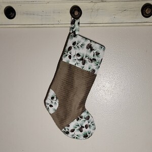 Könnte beinhalten: Ein brauner und weißer Weihnachtsstrumpf mit einem Tannenzapfenmuster. Der Strumpf hängt an einem Haken an einer weißen Wand.